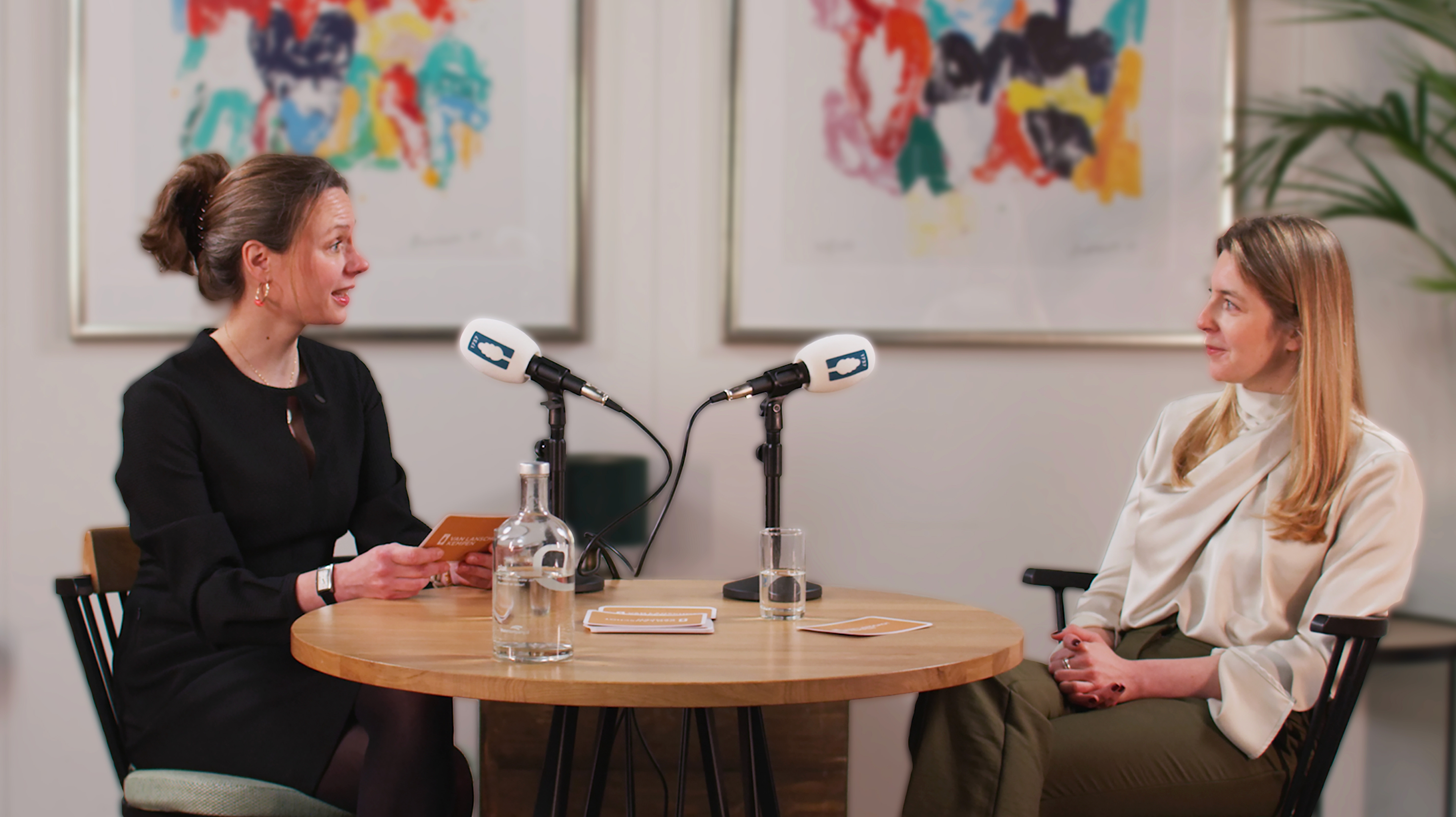 Two women sitting at a table with microphones and recording a podcast together.